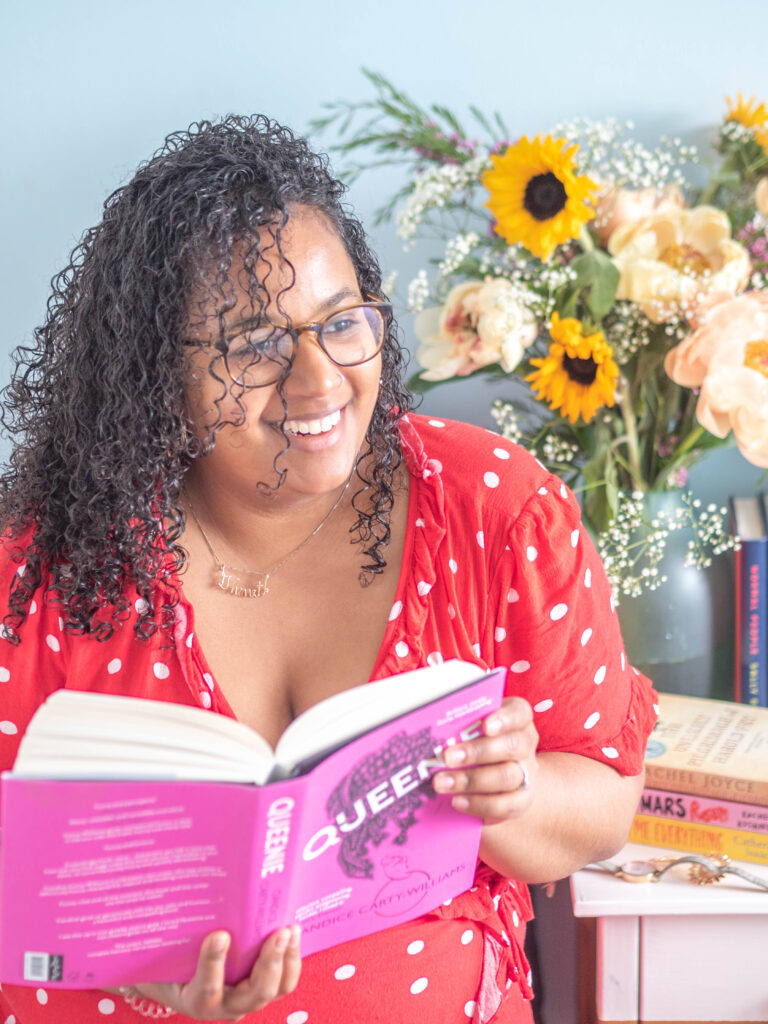 Ghenet holds a book and sits with it, several more books on display on the bedside table next to her.